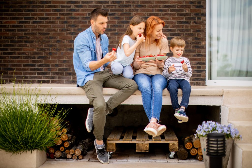 Family with a mother, father, son and daughter sitting outside on a steps of a front porch of a brick house and eating strawberries